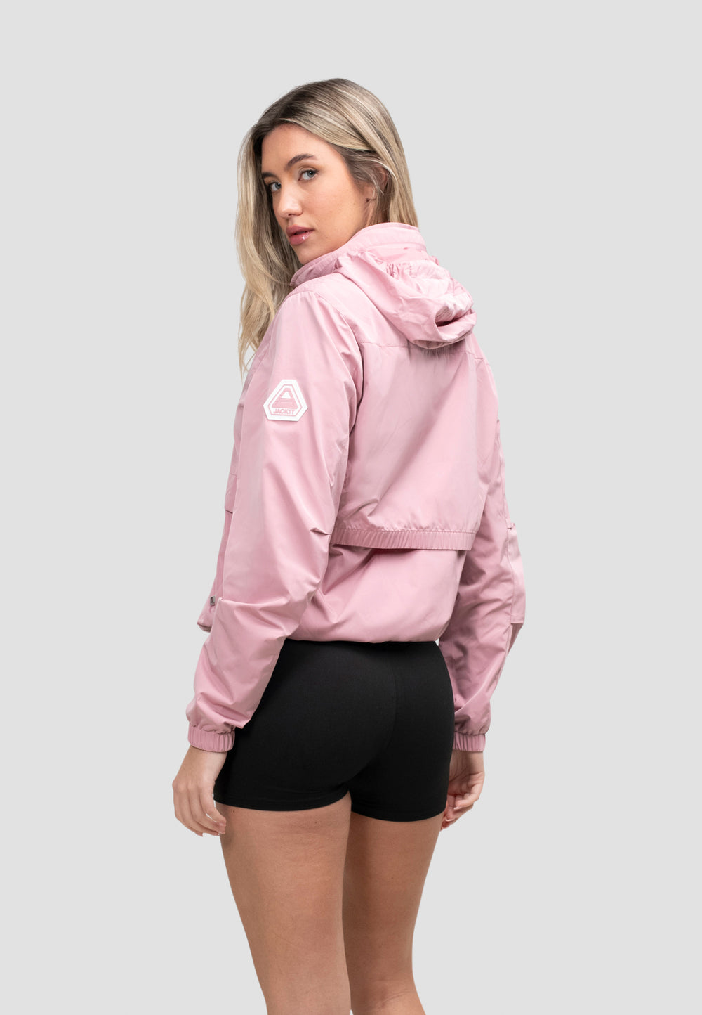 Model wearing a women's pink windbreaker, showcasing the back view, featuring a stow-away hood and a lightweight design.