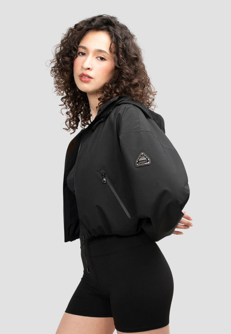 Ladies Cropped Black Jacket displayed on a model with curly hair, featuring an adjustable hood and zip pockets.