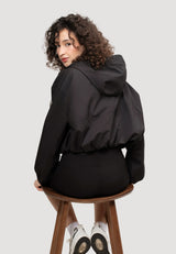 Back view of a woman wearing a lightweight Orpheus Cropped Jacket Black, showcasing a cropped fit and adjustable hood. Ladies Cropped Black Jacket.