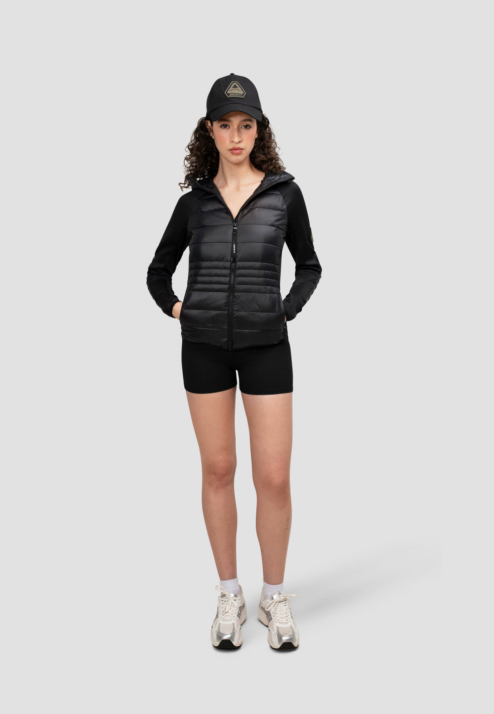 Model wearing a women's black hooded jacket, paired with black shorts and a cap, suitable for transitional weather.
