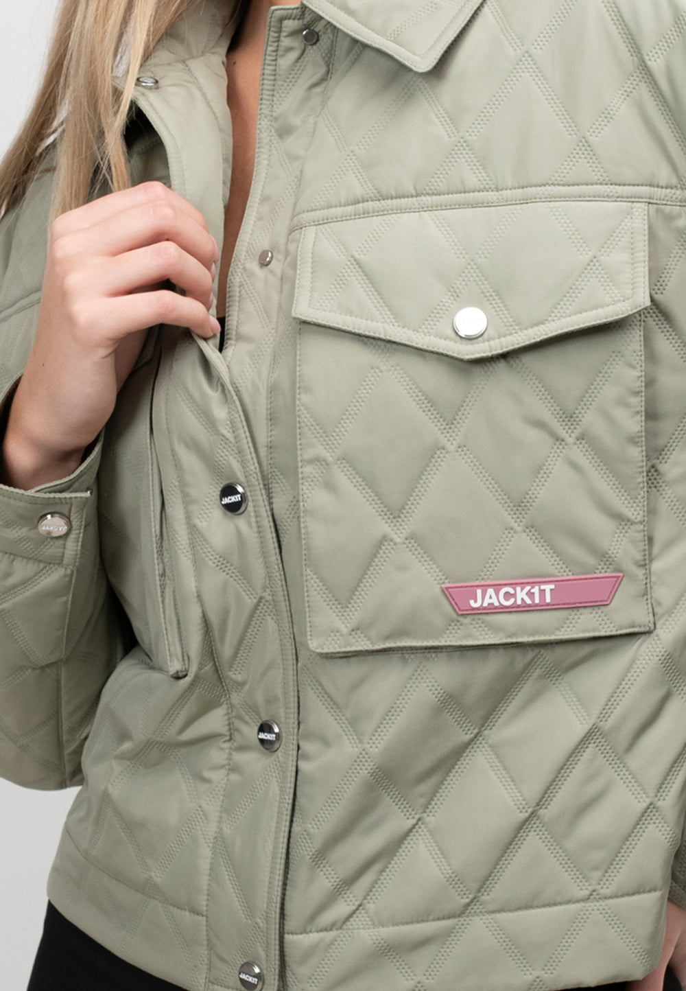 Close-up of a women's sage quilted shirt jacket featuring a buttoned pocket and brand logo detail.