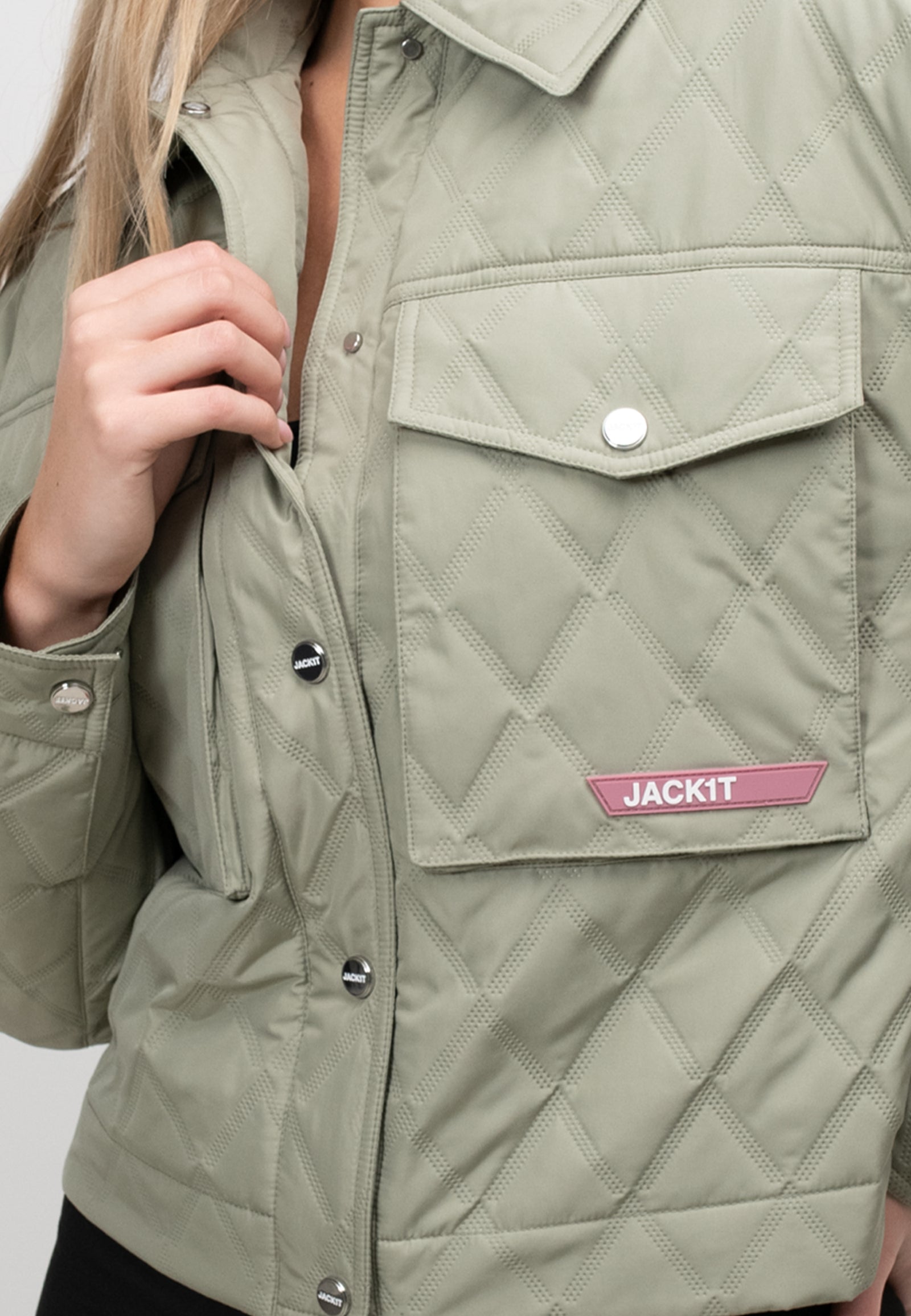 Close-up of a women's sage quilted shirt jacket featuring a buttoned pocket and brand logo detail.