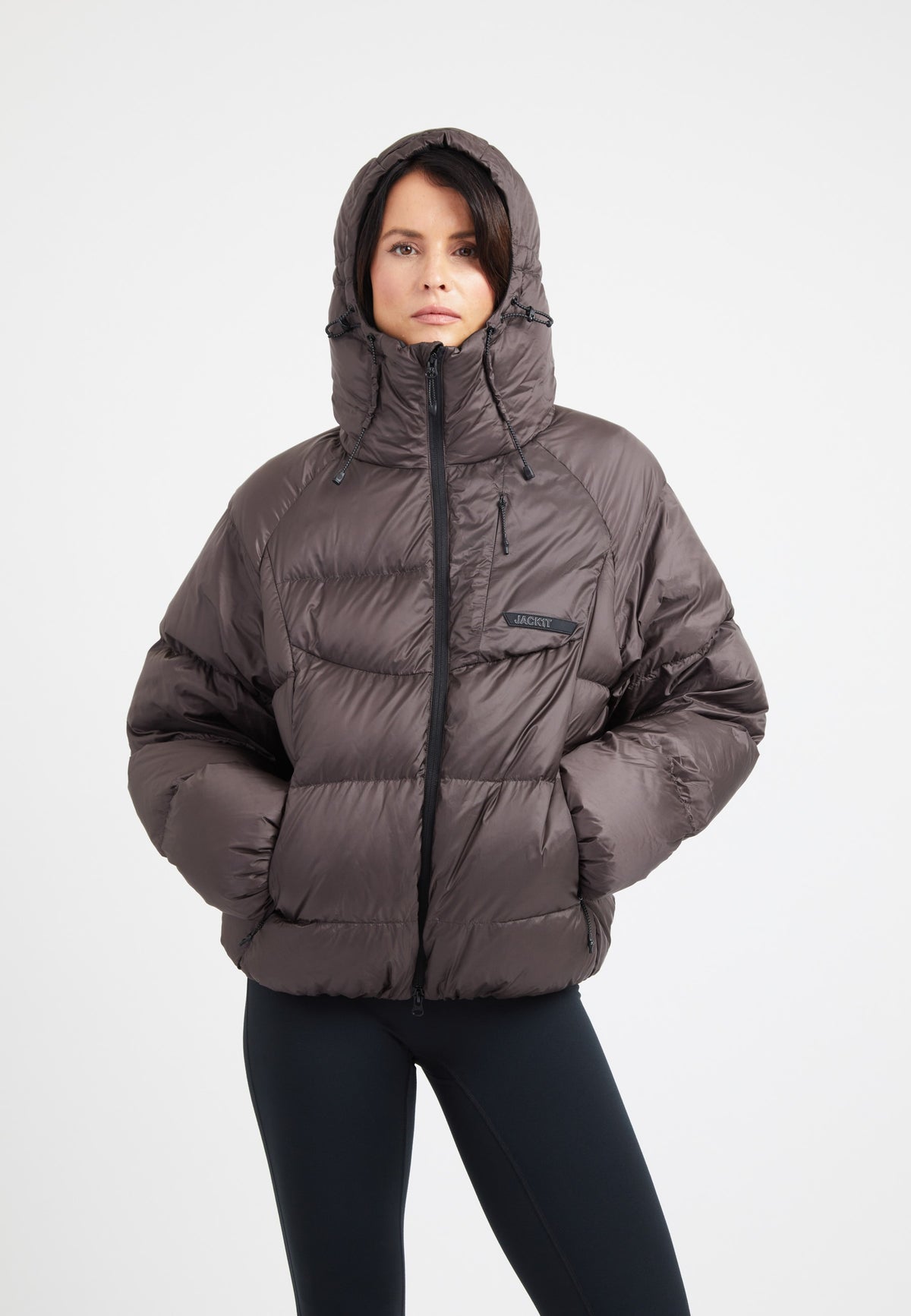 Woman wearing an oversized puffer brown jacket with a hood and zip pockets, showcasing its modern silhouette and down fill.