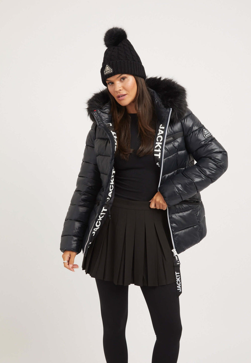 Model wearing a black womens puffer jacket with faux-fur trim and reflective badge, paired with black skirt and beanie.